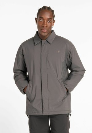 Junge Person mit geflochtenem Haar, die eine graue New Balance Jacke trägt und die Hände in den Taschen hat, stehend vor einem schlichten weißen Hintergrund.