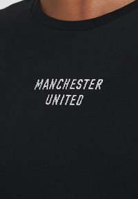 Czarna, krótko rękawowa koszulka z białym haftowanym napisem "MANCHESTER UNITED" na piersi. Gładki materiał bawełniany i okrągły dekolt.