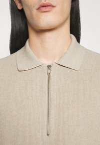 Gros plan d'une personne portant un pull beige en maille zippé avec un col, montrant les lèvres, le cou et les cheveux lisses et foncés.
