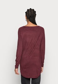 Bordeauxfarbener Langarmtop mit Rundhalsausschnitt, strukturiertem Stoff und lässiger Passform, mit abgerundetem Saum. Die Haare des Models sind zu Zöpfen geflochten.