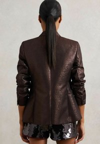 Hnědý kovový blejzr s výraznými rameny, úzkými rukávy a rozšířeným spodním lemem; v kombinaci s flitrovými šortkami v různých odstínech šedé.