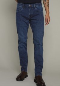 Džínové kalhoty v tmavě modré barvě, rovného střihu, s pěti kapsami, poutky na opasek a předním zapínáním na knoflík. Nošeny s hnědými kotníkovými botami.