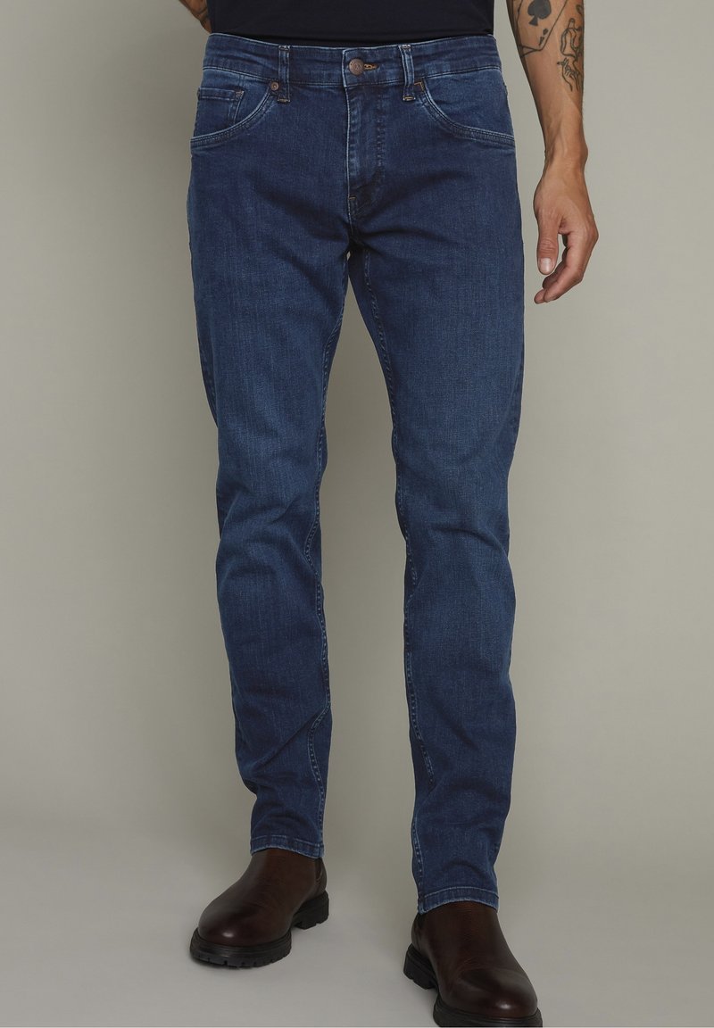 Džínové kalhoty v tmavě modré barvě, rovného střihu, s pěti kapsami, poutky na opasek a předním zapínáním na knoflík. Nošeny s hnědými kotníkovými botami.