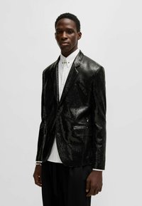 Schwarzer Pailletten-Blazer mit Reverskragen, einreihiger Knopfverschluss, zwei Seitentaschen, getragen über einem weißen Hemd, kombiniert mit schwarzen Hosen.