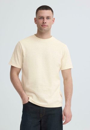 Mann med kort brunt hår iført en teksturert beige kortermet skjorte og mørke jeans, stående med en hånd i lommen mot en ensfarget bakgrunn.