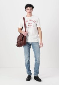 Brun lærveske med frontklaff, båret av en modell i en hvit bomulls-T-skjorte og lyseblå jeans, stående mot en ensfarget bakgrunn.