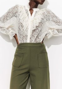 Blouse en dentelle blanche avec un col et des poignets volants, associée à un pantalon taille haute olive avec deux poches avant.