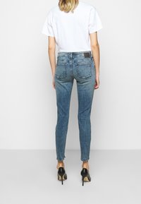 Mujer de pie con la espalda vuelta, vistiendo una camiseta blanca y jeans ajustados azules con tacones altos negros, fondo minimalista.