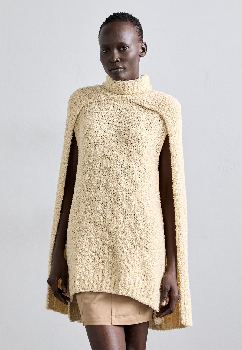 Beige knitted cape with a high collar, featuring a textured, fuzzy fabric. The cape drapes over the shoulders with side slits.