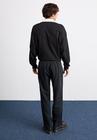 Sort langærmet bluse med hvid krave, parret med mørke bukser. Materialet ser blødt ud, med en afslappet pasform og elastisk talje. Sorte sko vist.