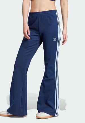 Osoba nosząca granatowe dresy Adidas z jasnoniebieskimi paskami po bokach i białe trampki, stojąca na gładkim tle.