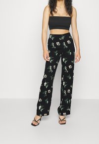 Žena stojí v černém topu bez ramínek a černých širokých kalhotách s zeleným a růžov ým květinovým vzorem, doplněná černými sandály na podpatku.