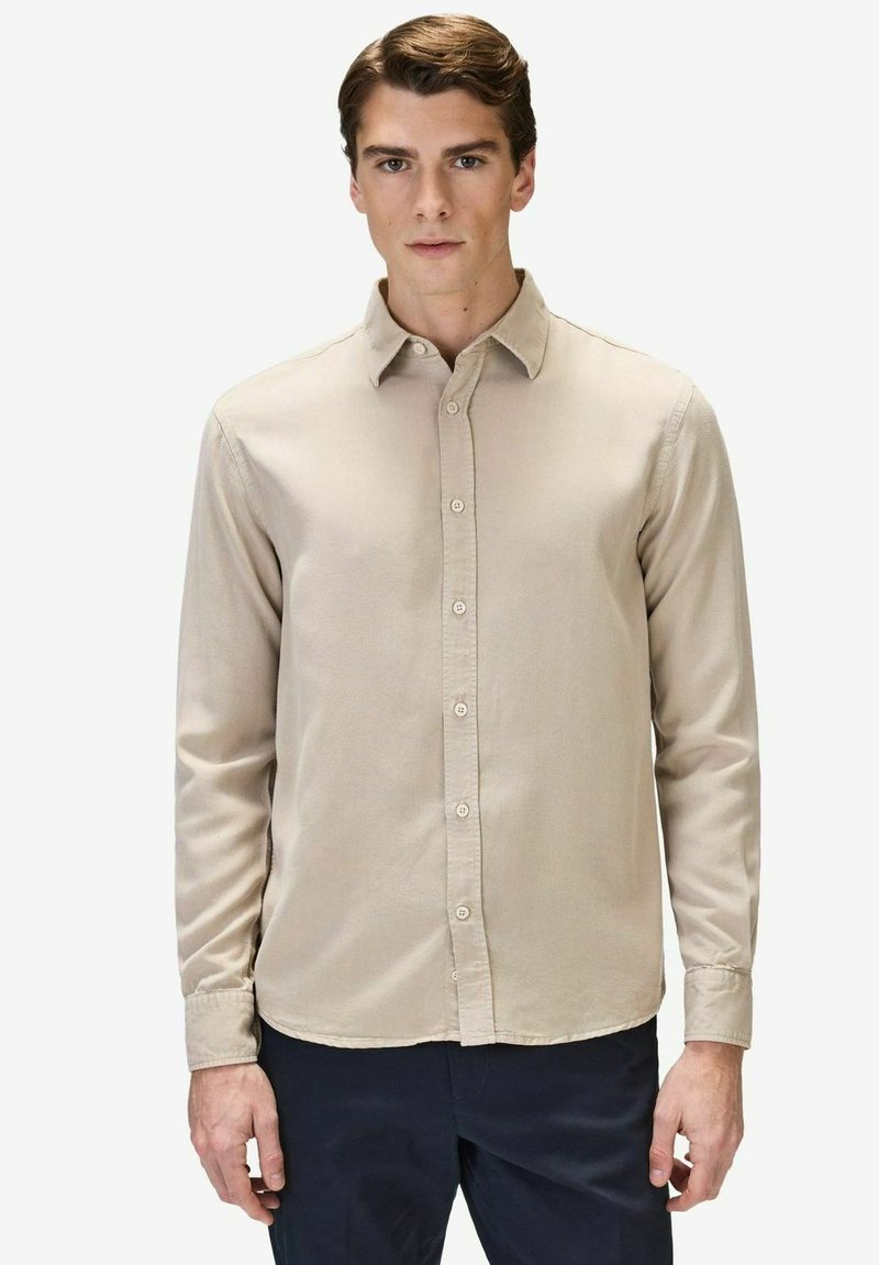 Beige napapaita, jossa on klassinen kaulus ja pitkät hihat, sujuvalla pinnalla ja suoran helmalla. Yhdistetty tummiin housuihin.