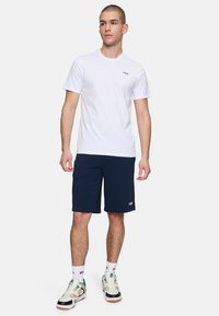Vit bomull t-shirt med en liten FILA-logotyp, tillsammans med marinblå shorts och vita sneakers med gröna och svarta accenter.