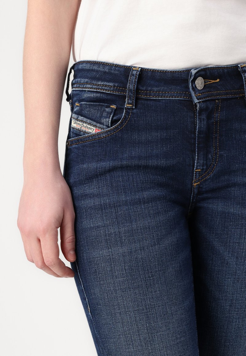 Dunkelblaue Jeans aus Denim mit taillierter Passform, ausgestattet mit gelben Nähten, einem Metallknopfverschluss und einem kleinen Label auf der Vorderseite der Tasche.