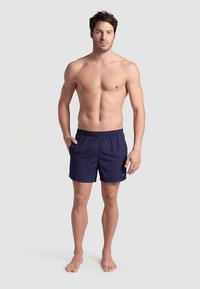 Mörkblå badshorts med elastisk midja och sidofickor, tillverkade av lättviktigt tyg, med en diskret logotyp på vänster lår.