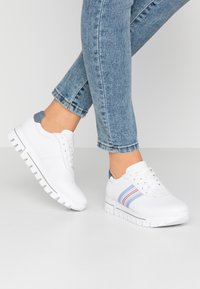 Scarpe sportive bianche con suola testurizzata, caratterizzate da un accento a strisce blu e rosse sul lato. Realizzate in materiale sintetico liscio.