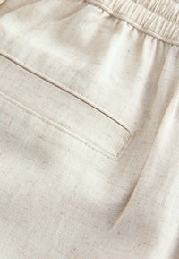 Gros plan sur un short en tissu beige montrant une ceinture élastique et une poche arrière cousue avec une texture en toile unie.