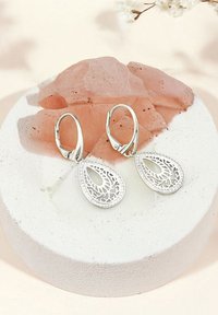 Silvriga tårformade örhängen med intrikata utskärningar, med en glänsande yta och låsning med fjäder, visade på en texturerad rund bas.