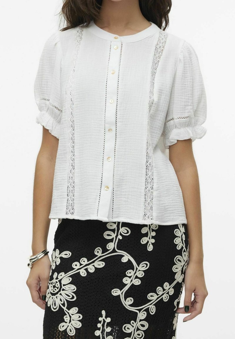 Femme portant un chemisier blanc texturé avec des détails en dentelle et des manches bouffantes, associé à une jupe noire avec une broderie florale crème.