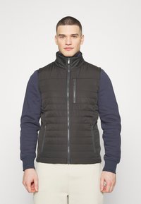 Černá vatovaná vesta s vysokým límcem, předním zipem a bočními kapsami, s prošívanými detaily a hladkou texturou látky.