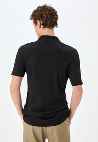 Polo negro de canalé con cuello y mangas cortas, que muestra una tela texturizada. Se lleva con pantalones beige, visto desde la espalda.