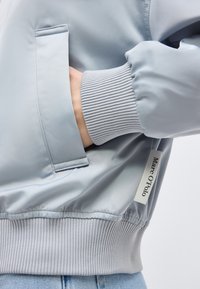 Käsi vaaleanharmaan takin taskussa, jossa on resorireunukset hihansuissa ja helmassa, näkyvä Marc O’Polo -merkkinen lappu sekä vaaleansiniset farkut.