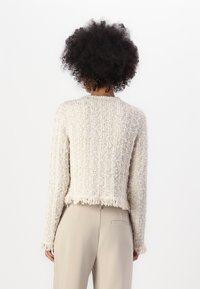Maglione lavorato a maglia color crema con un motivo testurizzato, frange all'orlo e alle maniche, abbinato a pantaloni beige chiaro.