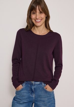Femme souriante portant un pull violet à manches longues et un jean bleu, les mains dans les poches, sur un fond clair uni.