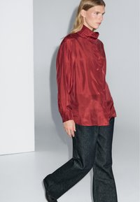 Red satin button-up shirt with a high collar, paired with dark denim wide-leg trousers. Smooth texture and relaxed fit.