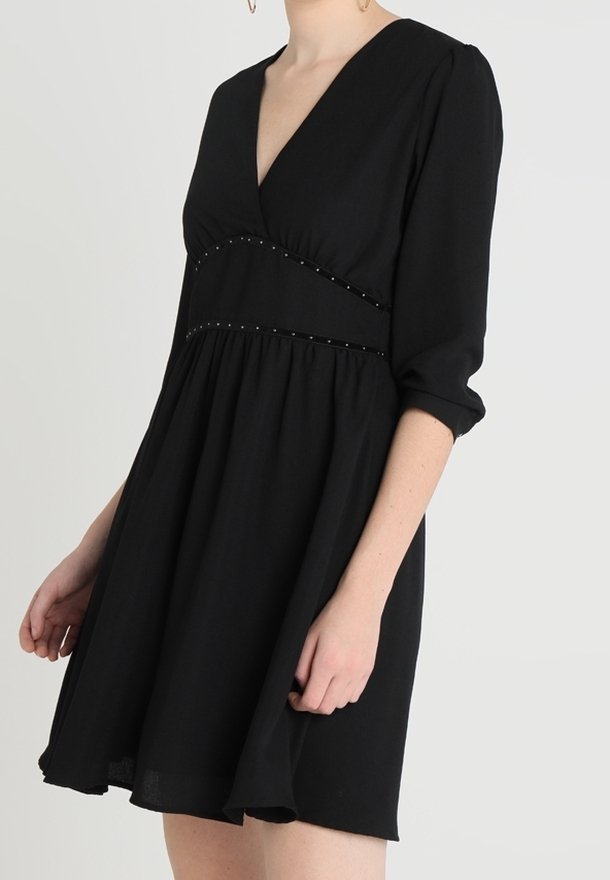Robe courte noire avec un décolleté en V profond, des manches trois-quarts, une taille cintrée ornée de petits clous, et une jupe évasée.