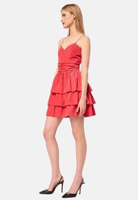 Abito rosso a strati con spalline sottili, vita arricciata e strati sfumati; abbinato a scarpe con tacco alto nere con accenti dorati.