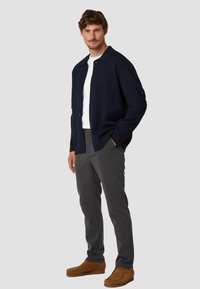 Marineblauwe button-up jas over een wit t-shirt, gecombineerd met grijze broeken en bruine schoenen. Eenvoudig ontwerp met een relaxed fit en minimale details.