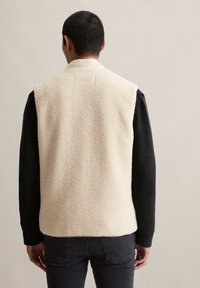 Crème-kleurige fleecevest met een gestructureerd oppervlak, voorzien van een hoge kraag en een zwart shirt met lange mouwen eronder. De achterkant toont geen zichtbare sluitingen.