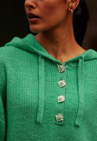 Roheline kudumiga kapuutsiga sviiter, millel on soonikstruktuur, nööriga ja kolm dekoratiivset hõbedast lillekest nööbina kaelaosas.