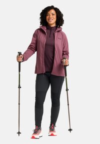 Una donna in giacca softshell bordeaux sopra un top scuro, pantaloni neri e scarpe da trekking multicolori, che tiene in mano dei bastoni da trekking.