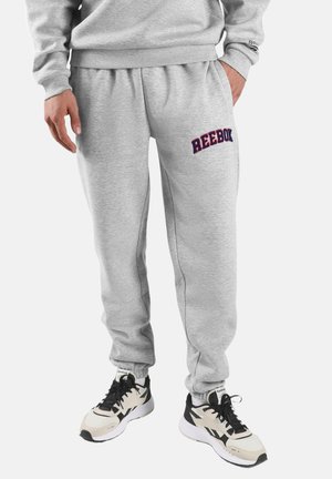 Man draagt lichtgrijze Reebok joggingbroek en bijpassend sweatshirt met witte en zwarte Reebok sneakers, staat met één hand in de zak.