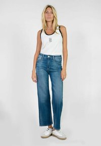 Top sin mangas blanco con ribetes negros y logo, combinado con jeans de pierna ancha de un azul claro y zapatillas blancas con suelas marrones, todo en un fondo liso.