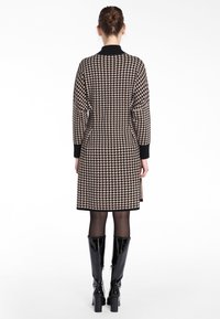 Robe longueur genoux avec un motif pied-de-poule, dotée d'un col et de poignets noirs, fabriquée à partir d'un tissu texturé ; associée à des bottes noires brillantes jusqu'aux genoux.