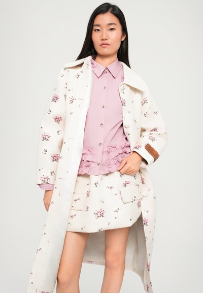 Fehér virágos trench coat rózsaszín virágokkal, világos rózsaszín fodros felsőre és hozzá illő szoknyára húzva, gombos díszítéssel és zsebekkel.