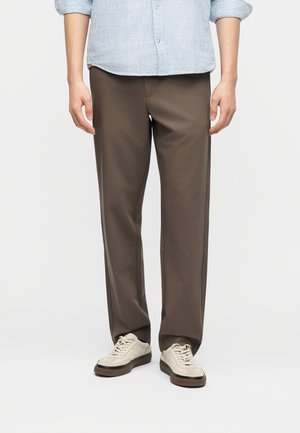 Άνδρας που φοράει καφέ παντελόνι κανονικής γραμμής, ανοιχτό γαλάζιο πουκάμισο με ρίγες και μπεζ αθλητικά παπούτσια με μαύρες σόλες, στέκεται μπροστά σε λευκό φόντο.