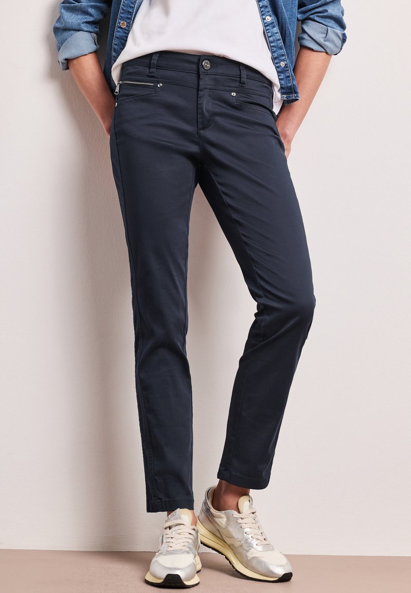 Personne portant un pantalon slim bleu marine, des baskets blanches, un haut blanc et une veste en jean retroussée, debout les mains dans les poches.