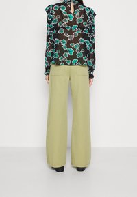 Blouse noire à motif floral avec des fleurs turquoise et roses, des manches à volants et un détail découpé dans le dos, associée à un pantalon large couleur olive clair.