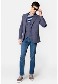 Granatowa marynarka w kratę z dopasowanym kamizelką na tle paskowanej koszuli, zestawiona z niebieskimi dżinsami i szarymi trampkami, dopełniona ciemnymi okularami przeciwsłonecznymi.