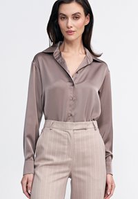 Koszula z długim rękawem zapinana na guziki w taupe satynie z kołnierzykiem typu spread. Połączona z jasnobeżowymi spodniami w pionowe paski. Gładka tekstura.