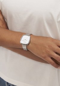 Orologio in metallo argentato con un quadrante rettangolare bianco, caratterizzato da semplici marker neri per le ore e un bracciale in metallo con maglie texturizzate, indossato su un polso.