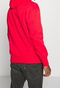 Felpa rossa in cotone con cappuccio regolabile, polsini a coste e orlo a coste, abbinata a jeans grigio scuro con dettaglio del logo sulla tasca posteriore.