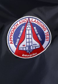 Ronde patch met een afbeelding van de ruimte Shuttle en de tekst "Approach and Landing Test", namen, en "Enterprise" op een donkere stofachtergrond.