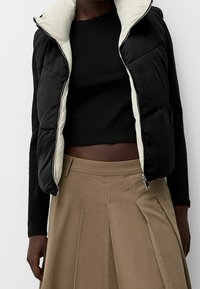 Black and cream puffer vest with a zipped front, featuring a high collar and patch pockets, paired with a fitted black top and beige pleated skirt.