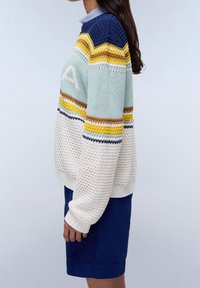 Kobieta ubrana w wielokolorowy, dziergany sweter w paski i niebieską spódnicę, stojąca bokiem na jasnym, jednolitym tle.